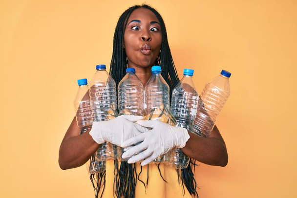 Mulher afro-americana com tranças segurando reciclagem garrafas de plástico fazendo cara de peixe com boca e olhos espremendo, louco e cômico.  - Foto, Imagem