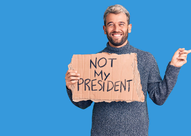Fiatal, jóképű szőke férfi, aki nem tartja az elnököm karton zászlóját mosolyogva, boldogan mutogatva, kezével és ujjával az oldalára.  - Fotó, kép