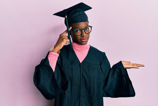 Young african american girl wearing graduation cap and ceremony robe confused and annoyed with open palm showing copy space and pointing finger to forehead. think about it.  - Foto, Bild