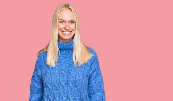 Mladá blondýna dívka na sobě vlny zimní svetr vypadá pozitivně a šťastně stojící a usmívá se s sebevědomým úsměvem ukazující zuby  - Fotografie, Obrázek