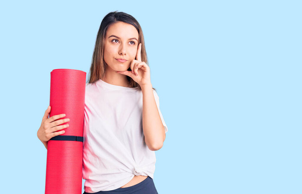 Young beautiful girl holding yoga mat serious face thinking about question with hand on chin, thoughtful about confusing idea  - 写真・画像