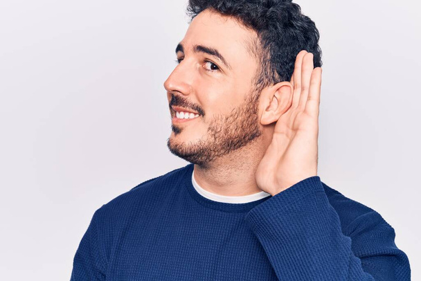 Young hispanic man wearing casual clothes smiling with hand over ear listening and hearing to rumor or gossip. deafness concept.  - Photo, Image
