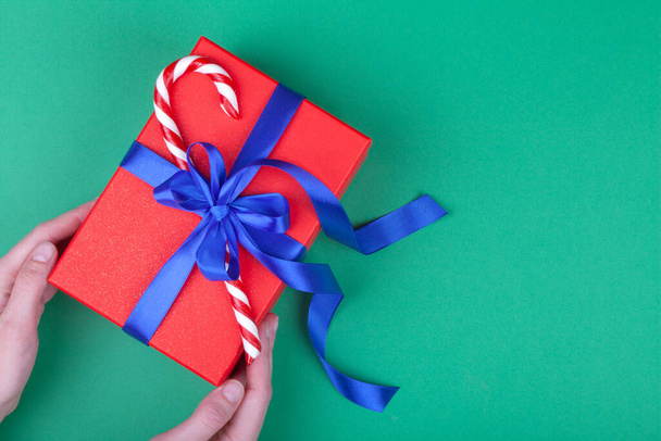 woman hands holding red gift with blue bow and Christmas candy on green background. Flat lay. Copy space. Minimal composition. - Valokuva, kuva
