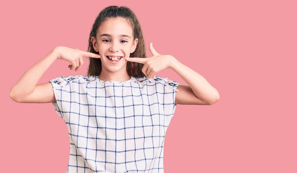 Мила дівчинка з іспаномовним дитячим одягом усміхається весело показуючи і вказуючи пальцями зуби і рот. концепція здоров'я зубів
.  - Фото, зображення