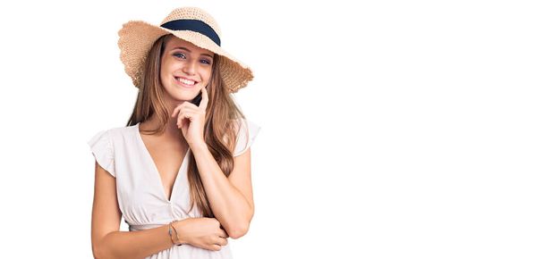 Young beautiful blonde woman wearing summer dress and hat looking confident at the camera with smile with crossed arms and hand raised on chin. thinking positive.  - Foto, Bild