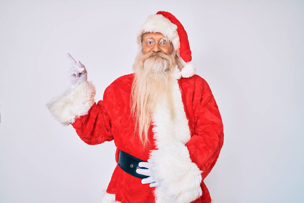 Alter älterer Mann mit grauen Haaren und langem Bart im Weihnachtsmann-Kostüm lächelt glücklich und zeigt mit Hand und Finger zur Seite  - Foto, Bild