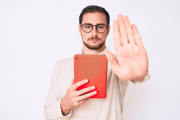 młody przystojny mężczyzna za pomocą touchpad urządzenie z otwartą ręką robi znak stop z poważnym i pewnym siebie wyrazem twarzy, gest obrony  - Zdjęcie, obraz