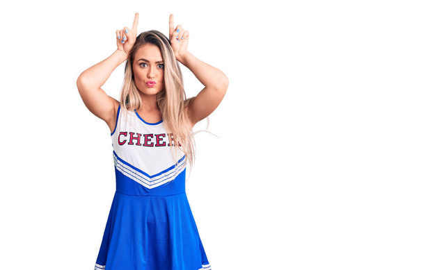 Giovane bella donna bionda indossa uniforme cheerleader facendo gesto divertente con il dito sopra la testa come corna di toro  - Foto, immagini