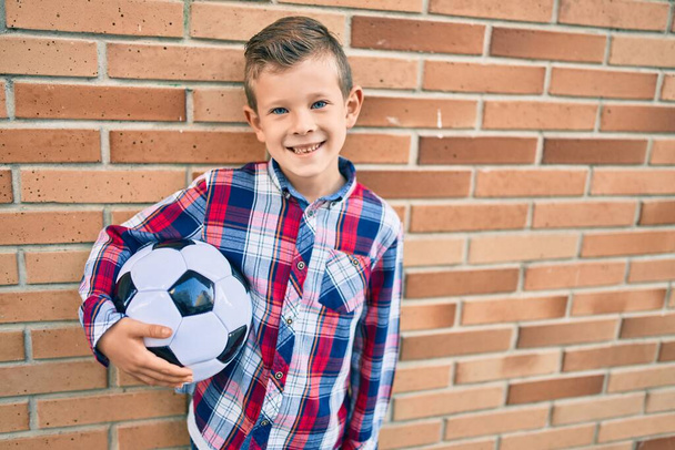 Очаровательный кавказский мальчик улыбается счастливо держа футбольный мяч, стоящий у города - Фото, изображение