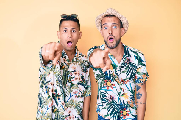 Junge schwule Paar von zwei Männern mit Sommermütze und hawaiianischem Hemd zeigt mit dem Finger überrascht voraus, offenen Mund erstaunte Miene, etwas auf der Vorderseite  - Foto, Bild