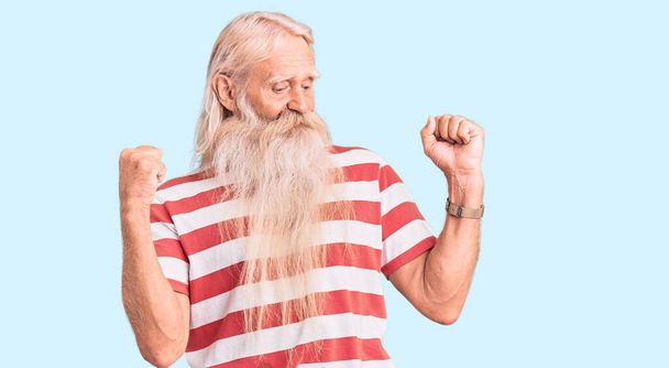 Alter älterer Mann mit grauen Haaren und langem Bart, der ein gestreiftes T-Shirt trägt, fröhlich und fröhlich tanzt, lässig lächelt und selbstbewusst Musik hört  - Foto, Bild