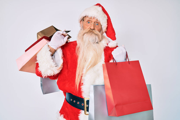Staruszek z siwymi włosami i długą brodą w kostiumie Świętego Mikołaja, trzymający torby na zakupy bez pojęcia i zdezorientowany wyraz twarzy. koncepcja wątpliwości.  - Zdjęcie, obraz