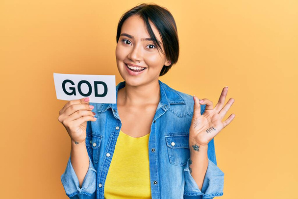 Όμορφη νεαρή γυναίκα με κοντά μαλλιά κρατώντας χαρτί με τη λέξη θεός κάνει εντάξει σημάδι με τα δάχτυλα, χαμογελώντας φιλικό gesturing εξαιρετικό σύμβολο  - Φωτογραφία, εικόνα
