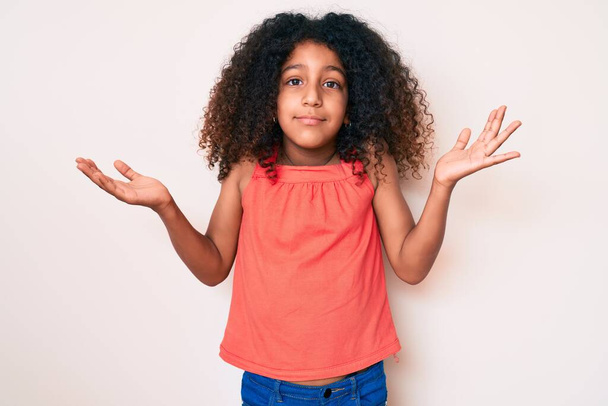 Criança afro-americana com cabelo encaracolado vestindo roupas casuais sem noção e confundidas com braços abertos, sem ideia e rosto duvidoso.  - Foto, Imagem