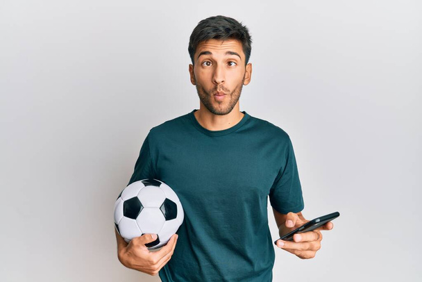 Молодой красивый мужчина, держащий футбольный мяч, глядя на приложение для ставок на смартфон, делает рыбное лицо с ртом и щурящимися глазами, сумасшедший и комичный.  - Фото, изображение