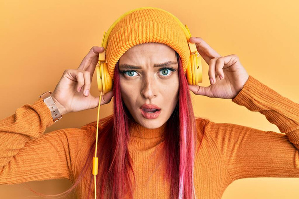 Young caucasian woman listening to music using headphones in shock face, looking skeptical and sarcastic, surprised with open mouth  - Photo, Image