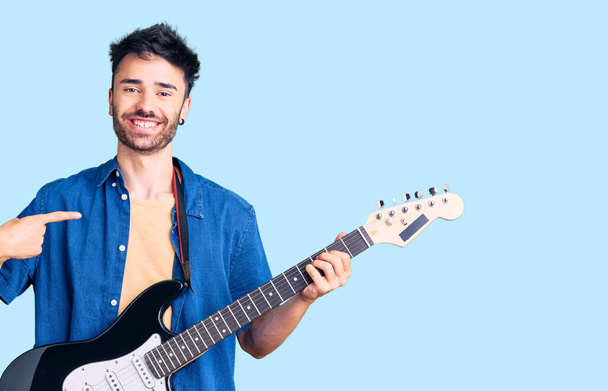 Giovane uomo ispanico suonare la chitarra elettrica puntando il dito verso un sé sorridente felice e orgoglioso  - Foto, immagini