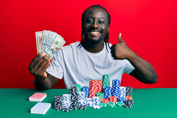 Poker oynayan yakışıklı genç bir adam elinde 100 dolarlık banknotlarla mutlu ve olumlu gülümsüyor, başparmağıyla mükemmel ve onay işareti yapıyor.  - Fotoğraf, Görsel