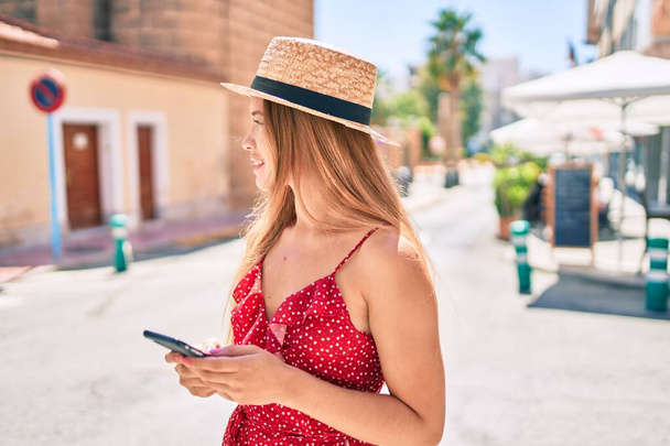 Νεαρό καυκάσιο τουριστικό κορίτσι χαμογελώντας χαρούμενος χρησιμοποιώντας smartphone στην πόλη. - Φωτογραφία, εικόνα