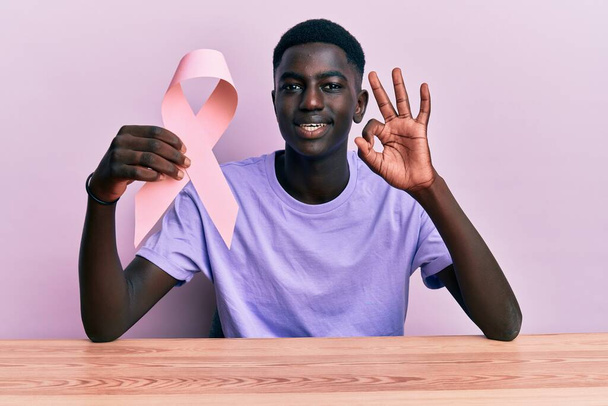 Young african american man holding pink cancer ribbon sitting on the table doing ok sign with fingers, smiling friendly gesturing excellent symbol  - Foto, afbeelding