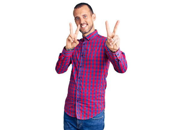 Ein junger gutaussehender Mann in lässiger Kleidung lächelt in die Kamera und zeigt Finger, die ein Siegeszeichen machen. Nummer zwei.  - Foto, Bild