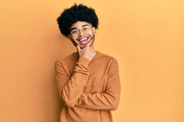 Junger afrikanisch-amerikanischer Mann mit Afro-Haaren in lässigem Winterpullover, der selbstbewusst in die Kamera lächelt, mit verschränkten Armen und erhobener Hand am Kinn. Positives Denken.  - Foto, Bild