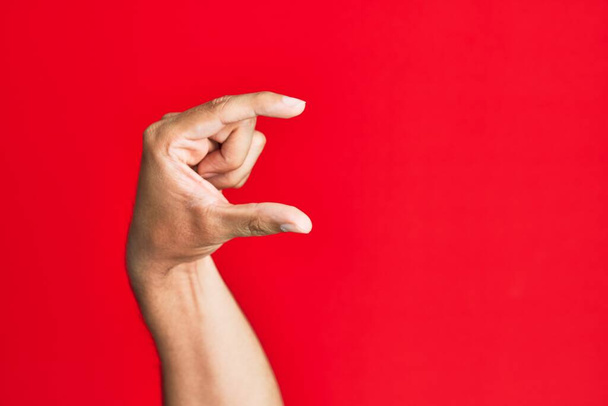 Valkoihoisen nuoren miehen käsivarsi punaisen, eristetyn taustan päällä ja näkymättömän asian ottaminen, sormilla varustettu esine, joka näyttää tilaa  - Valokuva, kuva