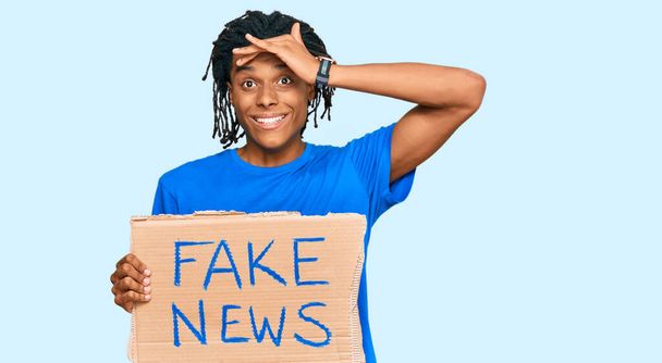 Mladý africký Američan drží falešné zprávy prapor zdůraznil a frustrovaný s rukou na hlavě, překvapený a rozzlobený obličej  - Fotografie, Obrázek