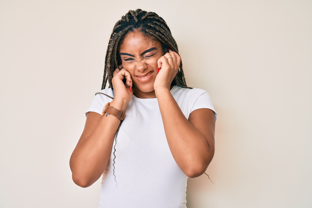 Młoda Afroamerykanka z warkoczami w białej koszuli zakrywającej uszy palcami z irytującym wyrazem twarzy dla hałasu głośnej muzyki. koncepcja głucha.  - Zdjęcie, obraz