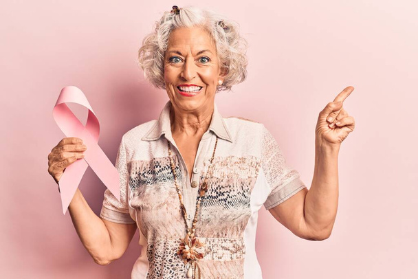 Ältere grauhaarige Frau mit rosa Krebsband lächelt glücklich und zeigt mit Hand und Finger zur Seite  - Foto, Bild