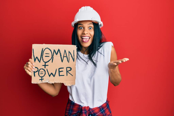 Junge afrikanisch-amerikanische Frau mit Architektenhut und Women-Power-Banner, die Errungenschaften mit glücklichem Lächeln und Siegerausdruck mit erhobener Hand feiert  - Foto, Bild