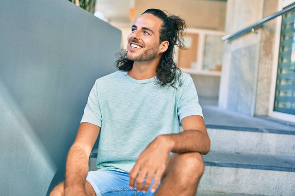 Νεαρός Ισπανός χαμογελάει χαρούμενος κοιτάζοντας στο πλάι και κάθεται στις σκάλες. - Φωτογραφία, εικόνα