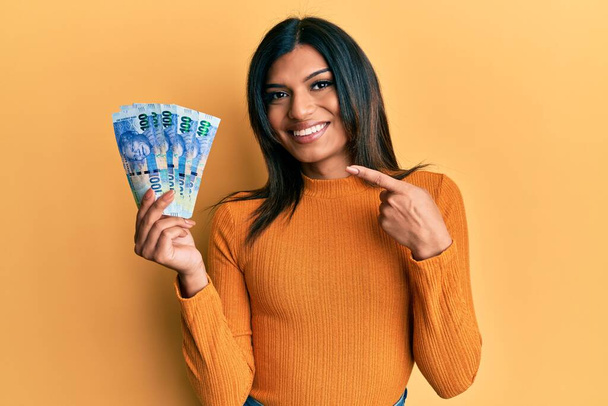 jong latijn transseksueel transgender vrouw houden zuid-afrikaanse 100 rand bankbiljetten glimlachen gelukkig wijzend met hand en vinger  - Foto, afbeelding
