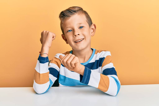 Roztomilé bělošské dítě v neformálním oblečení, které sedí na stole a ukazuje na zadní stranu se zdviženýma rukama a palci.  - Fotografie, Obrázek