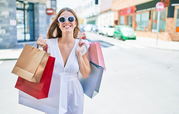 Joven mujer rubia sonriendo feliz sosteniendo bolsas de compras en la calle de la ciudad - Foto, Imagen