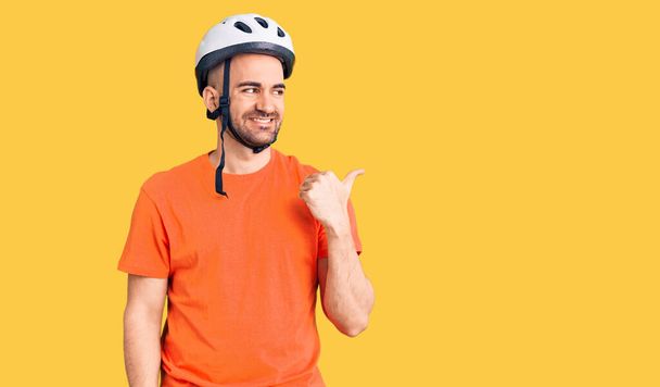 Young handsome man wearing bike helmet smiling with happy face looking and pointing to the side with thumb up.  - Fotó, kép
