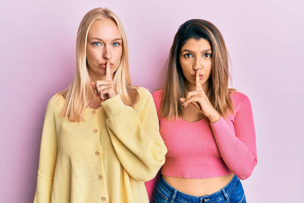 Zwei Freunde, die zusammen vor rosa Hintergrund stehen und mit dem Finger auf den Lippen um Ruhe bitten. Schweigen und geheimes Konzept.  - Foto, Bild