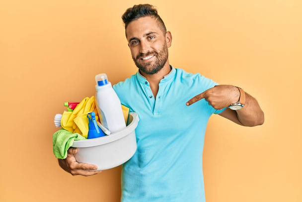 Hombre guapo con barba sosteniendo productos de limpieza señalando con el dedo a uno mismo sonriendo feliz y orgulloso  - Foto, imagen