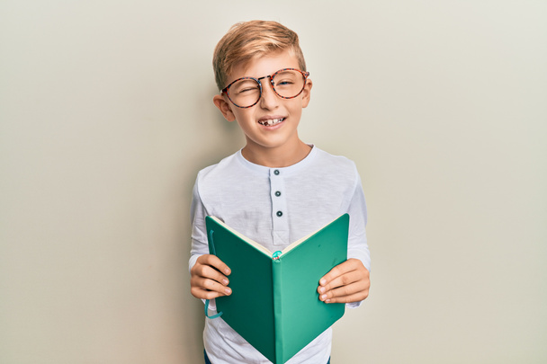 Klein blank jongetje dat een boek leest met een bril op knipogend kijkend naar de camera met sexy uitdrukking, vrolijk en vrolijk gezicht.  - Foto, afbeelding