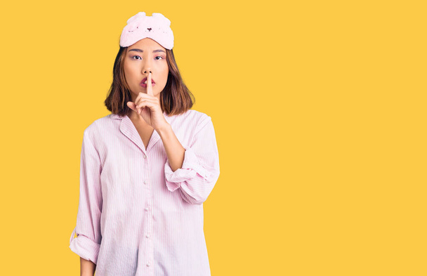 Menina chinesa bonita nova usando máscara de sono e pijama pedindo para ficar quieta com o dedo nos lábios. silêncio e conceito secreto.  - Foto, Imagem