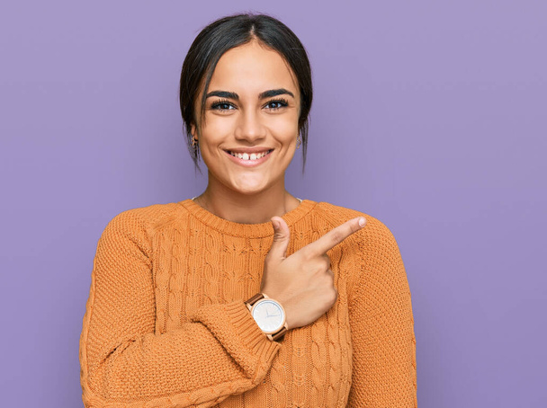 Νεαρή μελαχρινή γυναίκα που φοράει casual χειμωνιάτικο πουλόβερ χαρούμενη με χαμόγελο στο πρόσωπο δείχνοντας με το χέρι και το δάχτυλο στο πλάι με χαρούμενη και φυσική έκφραση στο πρόσωπο  - Φωτογραφία, εικόνα