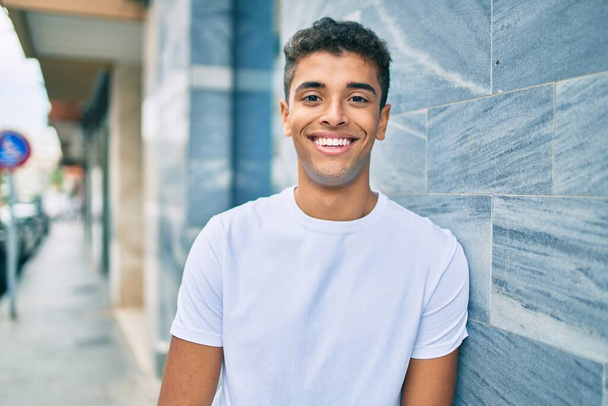Młody Latynos uśmiecha się szczęśliwy opierając się o ścianę w mieście. - Zdjęcie, obraz