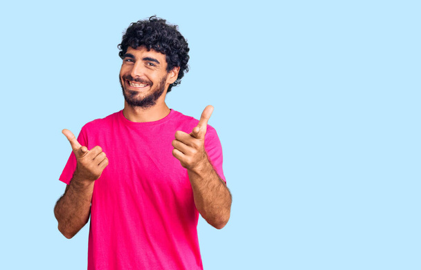 Schöner junger Mann mit lockigem Haar und Bär in lässigem rosafarbenem T-Shirt, der mit erhobenem Zeigefinger in die Kamera zeigt, mit fröhlichem und lustigem Gesicht. gute Energie und Stimmung.  - Foto, Bild