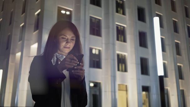 Woman alone holding a phone wearing black jacket and neck scarf, night shot  - Fotografie, Obrázek