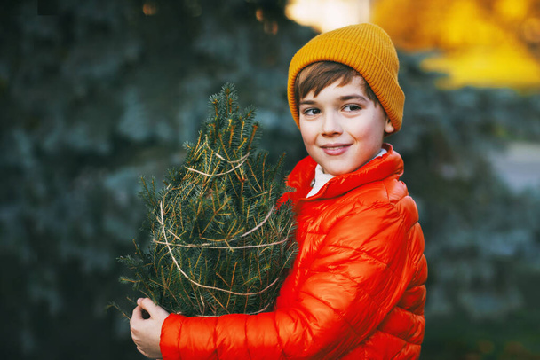 Een jongen in een fel oranje jasje en een gele hoed houdt een gekochte kerstboom in zijn handen, glimlacht en kijkt in de verte. Winkelen voor de vakantie. Voorbereiding van Kerstmis, Nieuwjaar - Foto, afbeelding