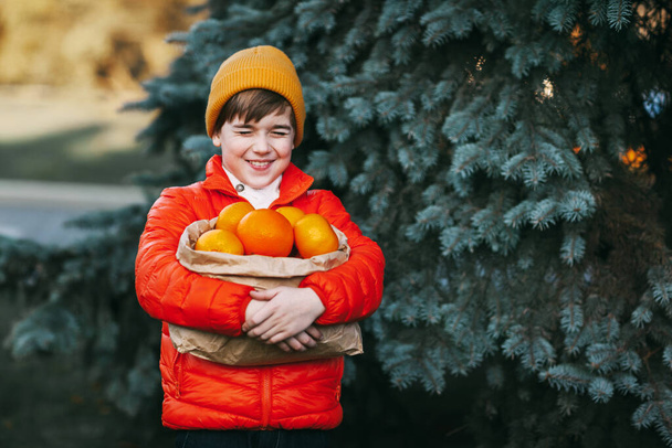 Un niño lindo con una chaqueta naranja brillante y un sombrero amarillo sostiene un paquete grande con naranjas en sus manos contra el fondo de un árbol de Navidad. Preparación para la Navidad, Año Nuevo - Foto, imagen