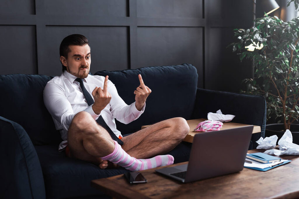 Angry businessman without pants showing rude gesture into the laptop's camera during online conference - Foto, imagen