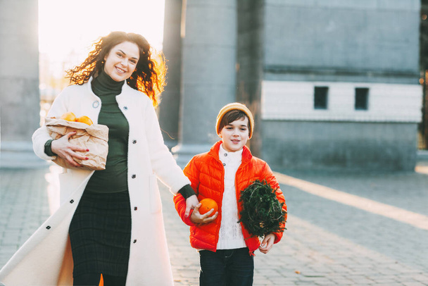 Een mooie moeder met haar zoon in een fel oranje jasje en een modieuze gele hoed lopen de winkel uit met een pakje sinaasappels en een kleine kerstboom. Winkelen voor de vakantie - Foto, afbeelding
