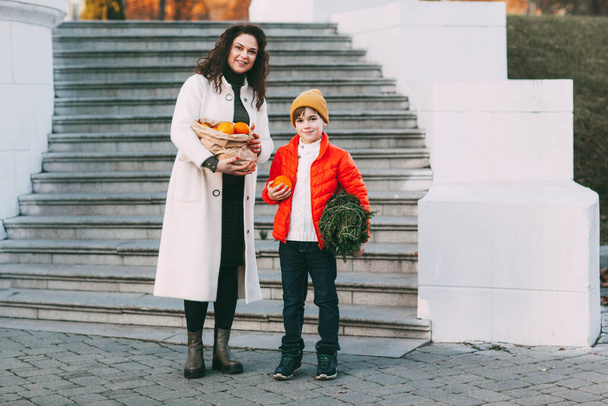 Parlak turuncu ceketli ve şık sarı şapkalı güzel bir anne elinde bir paket portakal ve küçük bir Noel ağacı ile mağazadan yürüyor. Tatil alışverişi. - Fotoğraf, Görsel