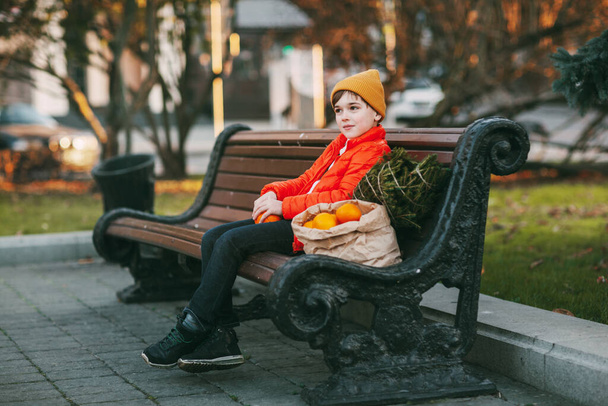 мальчик в ярко-оранжевой куртке и модной желтой шляпе сидит на скамейке, смеется, чистит апельсины, а рядом стоит дерево и большой мешок апельсинов. шоппинг на праздник. подготовка к - Фото, изображение